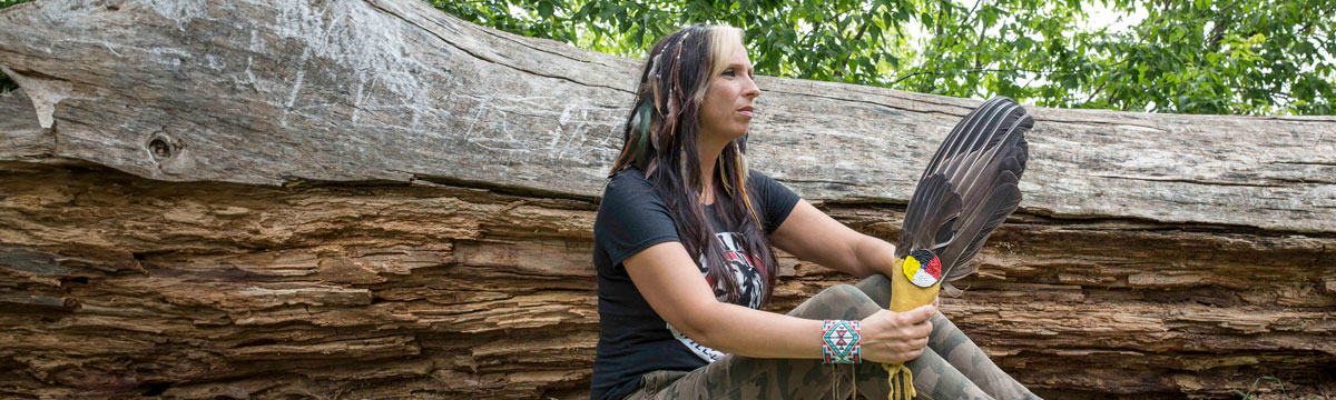 Pamela Palmater outside by a large tree, holding a bundle of eagle feathers, a beaded medicine wheel with red, black, white and yellow pinned below. 