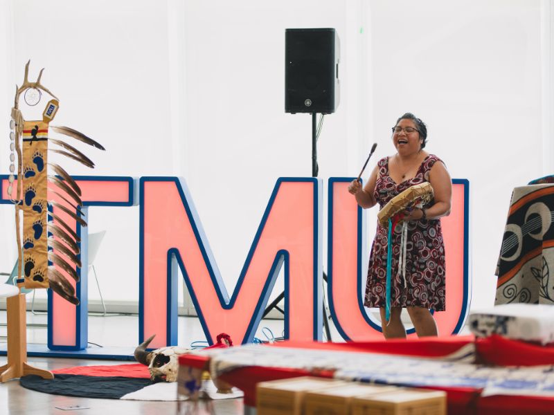 Amy Desjarlais sings and plays the hand drum, standing in front of large “TMU” letters and the TMU eagle staff.