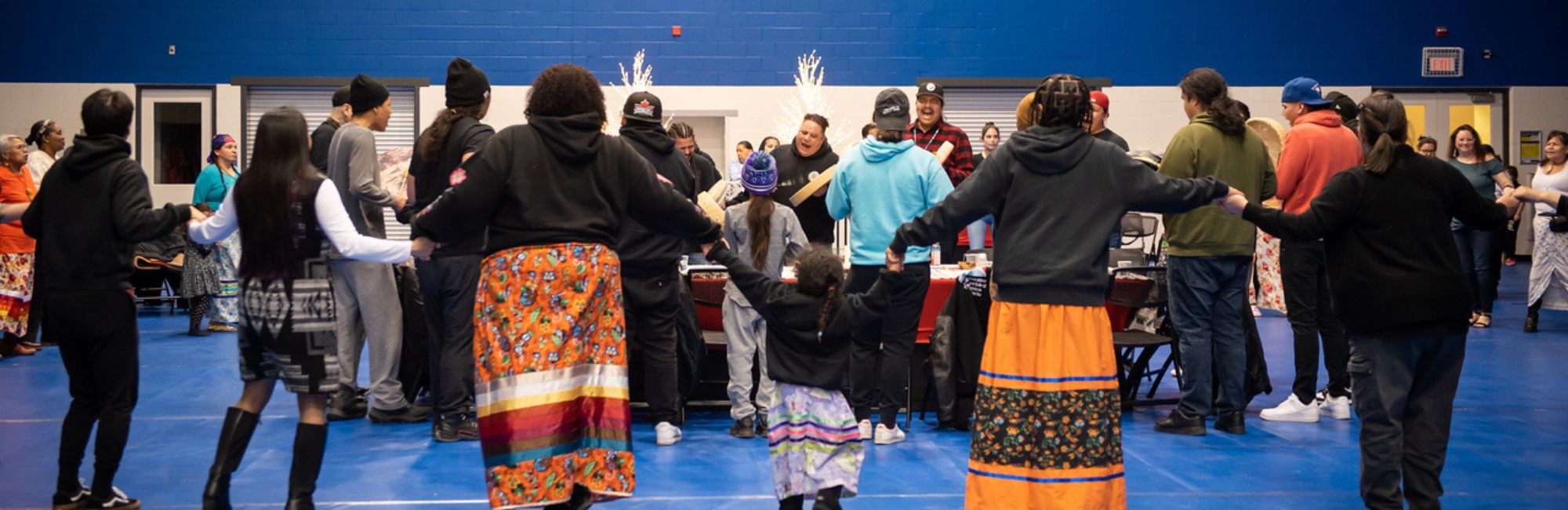 Community members hold hands in a circle at the round dance while others drum and sing. Some attendees are wearing ankle-length ribbon skirts.