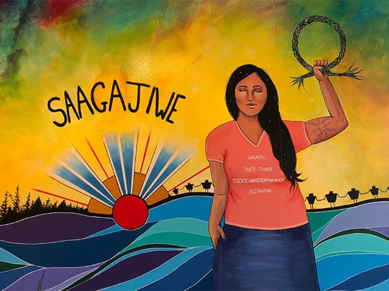  A mural featuring Indigenous art with a woman in the middle and a hill landscape behind her. On the left, the Saagajiwe logo is painted
