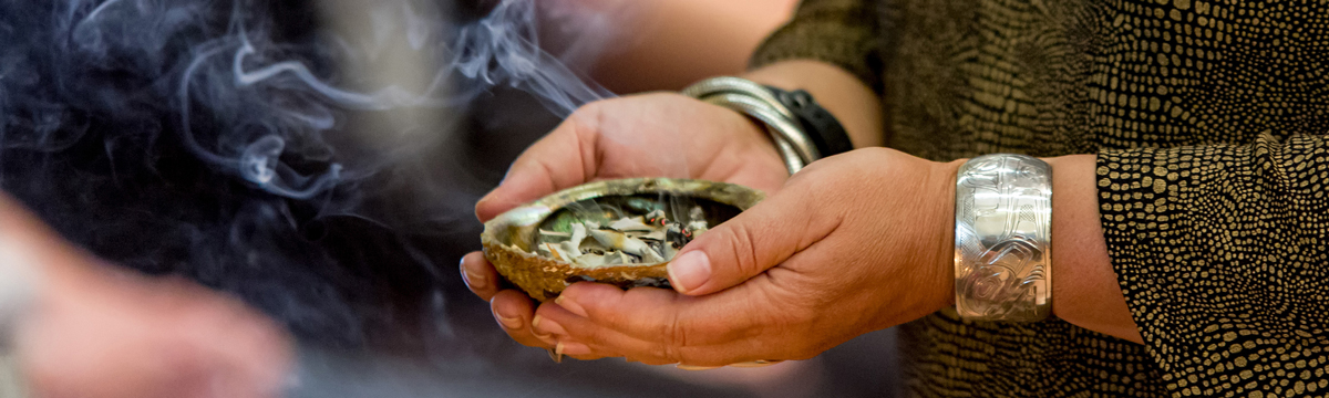 A person smudging: An abalone shell with sage leaves inside of it; smoke wafting above it