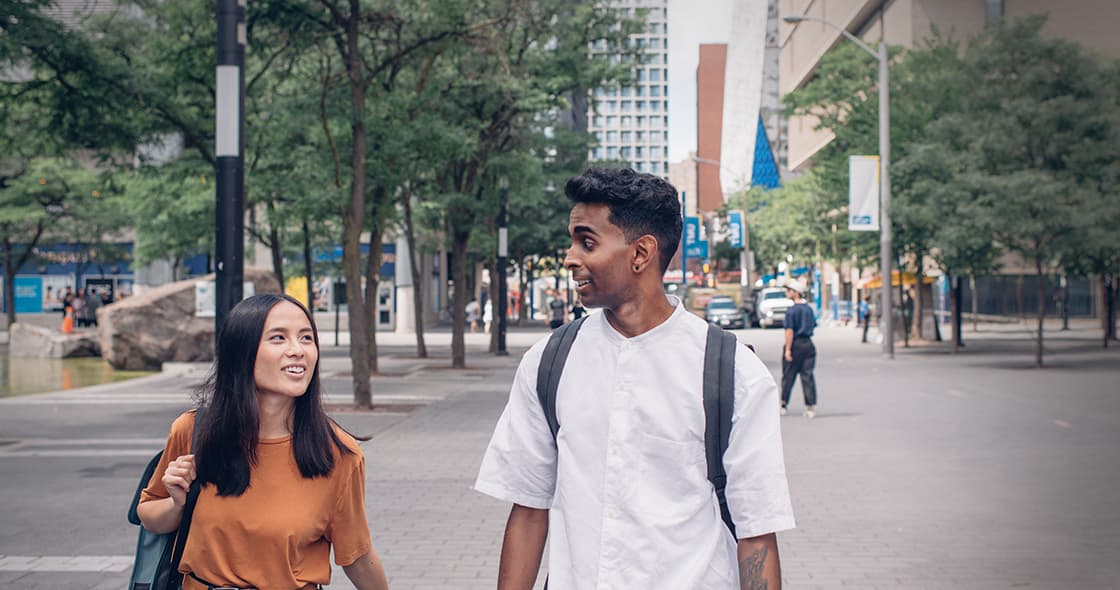 Two students walking on campus, looking at each other and speaking. Trees and Lake Devo can be seen in the background.