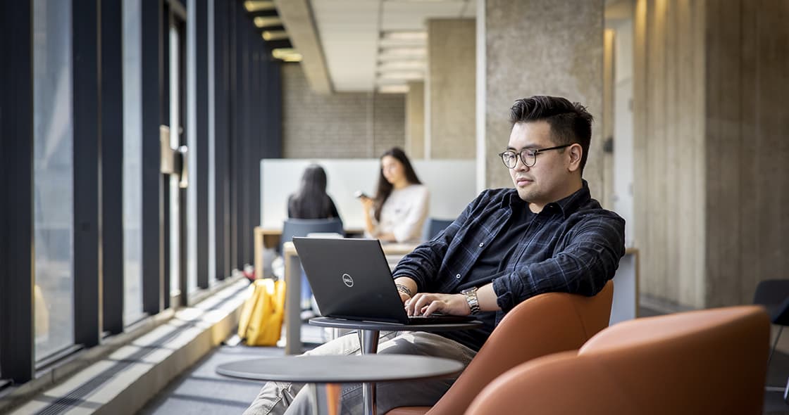 Student is working on his laptop in a TMU study lounge. Two students can be seen working in the background.