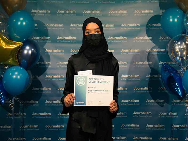 Layyan Mohamed-Birhan holds a certificate while standing in front of a blue backdrop that repeats, "Journalism at The Creative School."