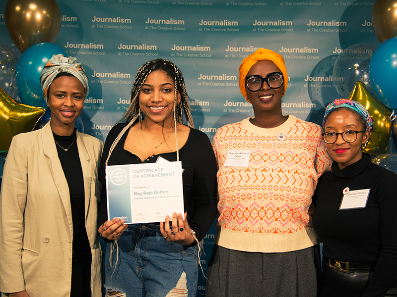 Ray Rojo Bolton holds a certificate while standing with Dahabo Ahmed Omer, Sagal Abdi and tatenda Mhaka in front of a blue backdrop that repeats, "Journalism at The Creative School."