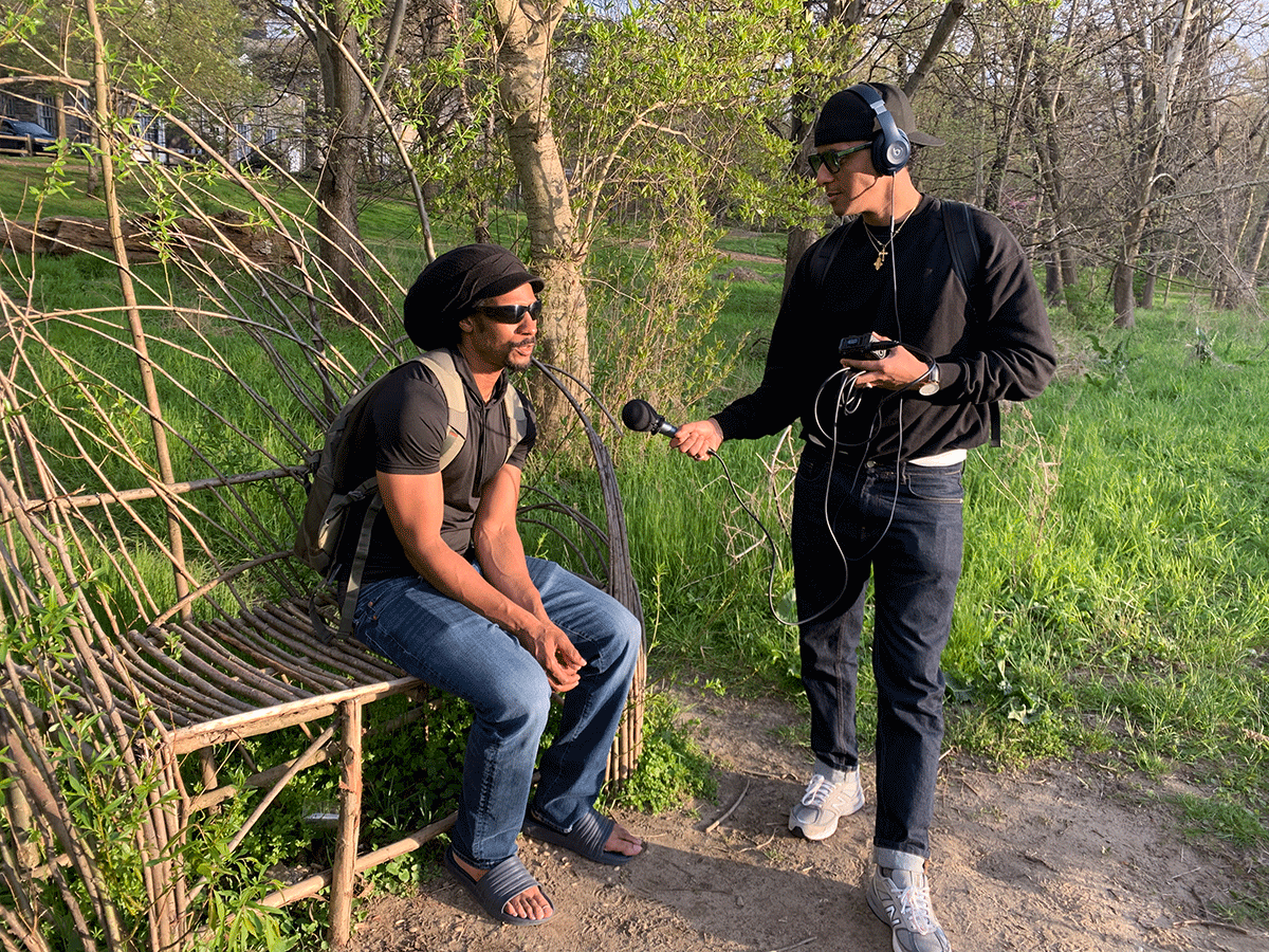 Amha standing with a microphone and recorder interviewing a subject who is sitting on a bench.