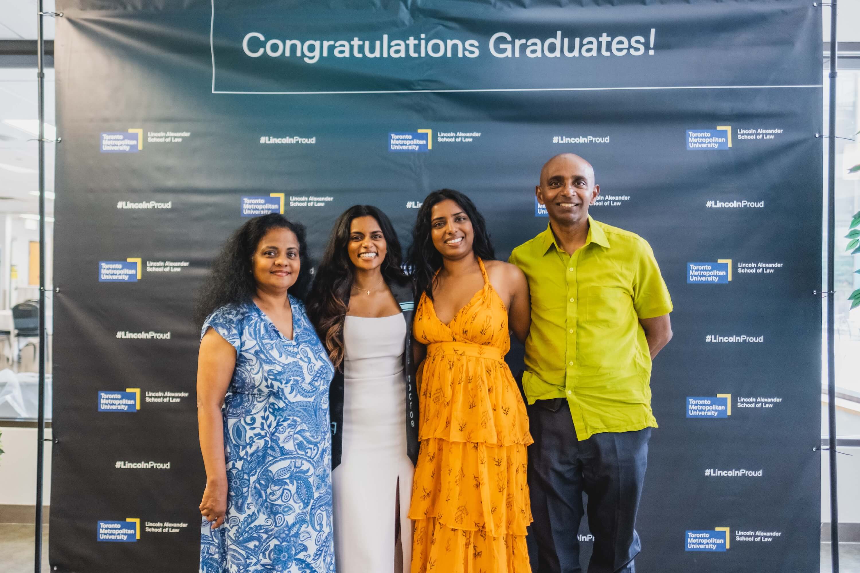 two young women, a man and a woman pose in front of a backdrop that says "Congratulations Graduates!"