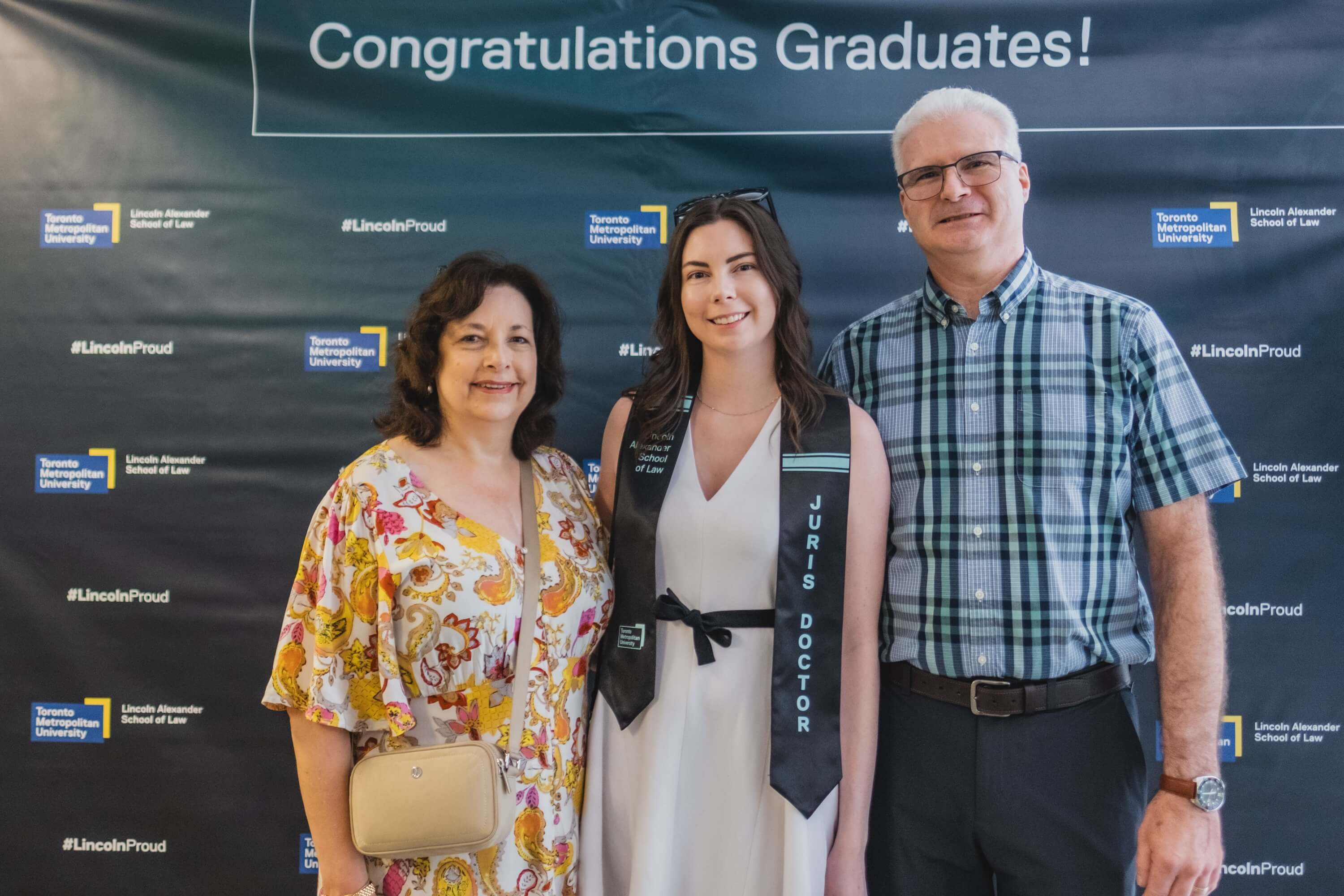 A young woman and an older man and woman pose for a photo in front of a backdrop that says "Congratulations Graduates!"