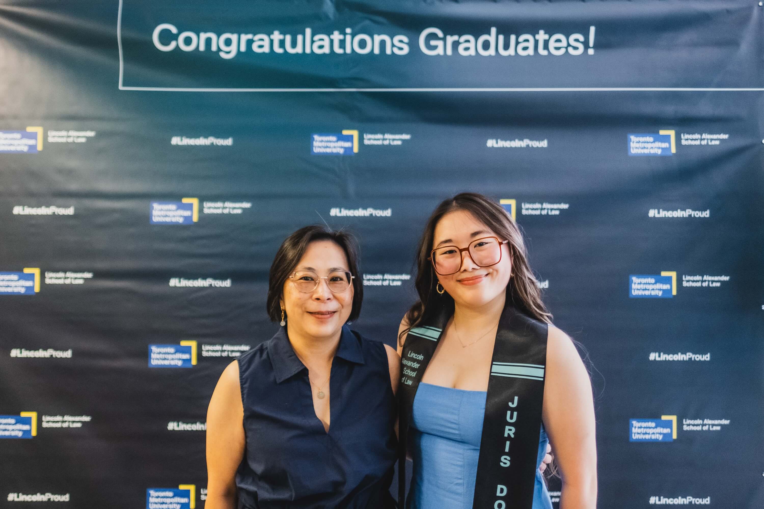 A young woman and a woman posing in front of a backdrop that says "Congratulations Graduate!"