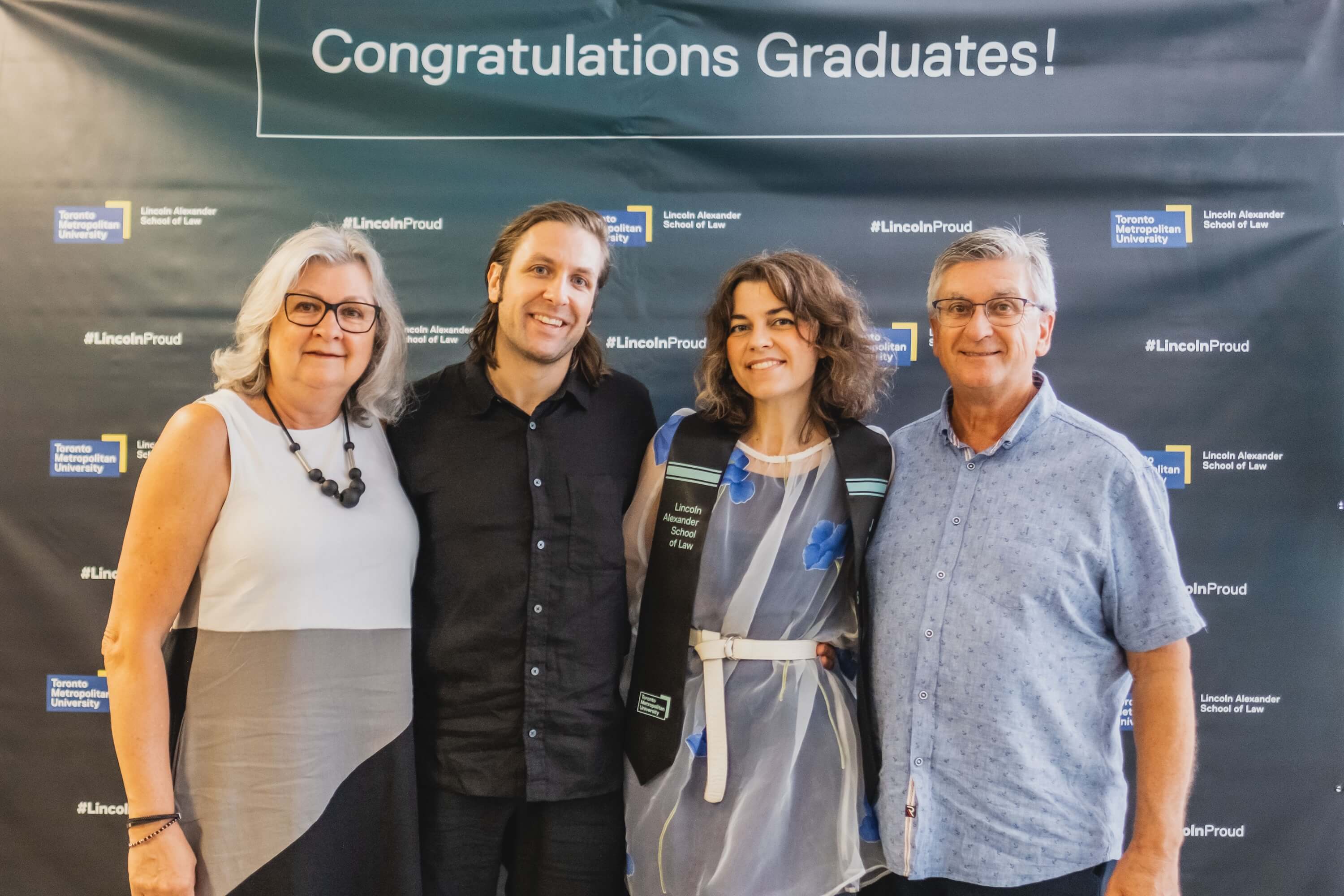Two men and two women, one a graduating student, pose for a photo in front of a backdrop that says "Congratulations Graduates!"