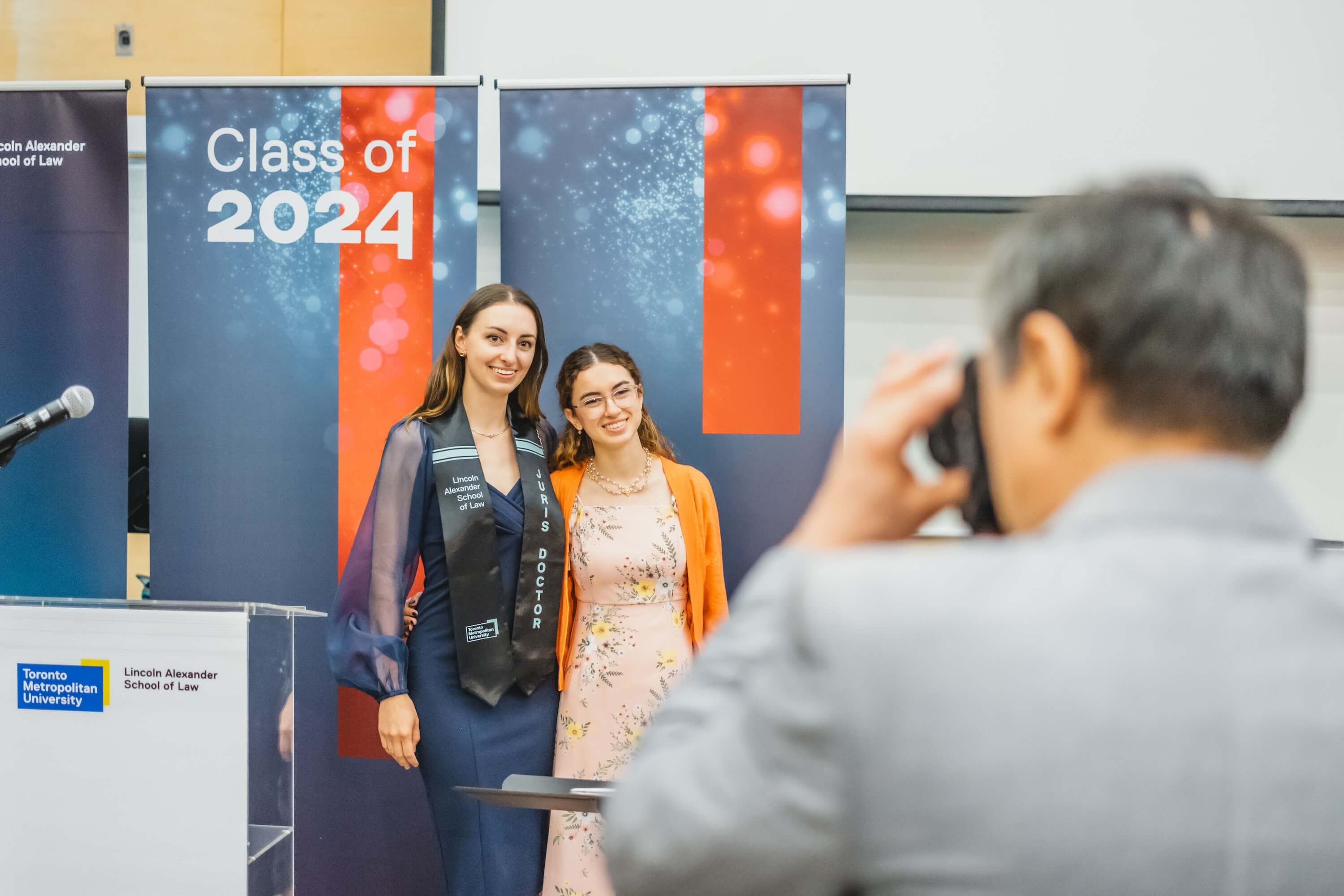 Two young women pose in front of a "Class of 2024" banner while an older man takes their photo.