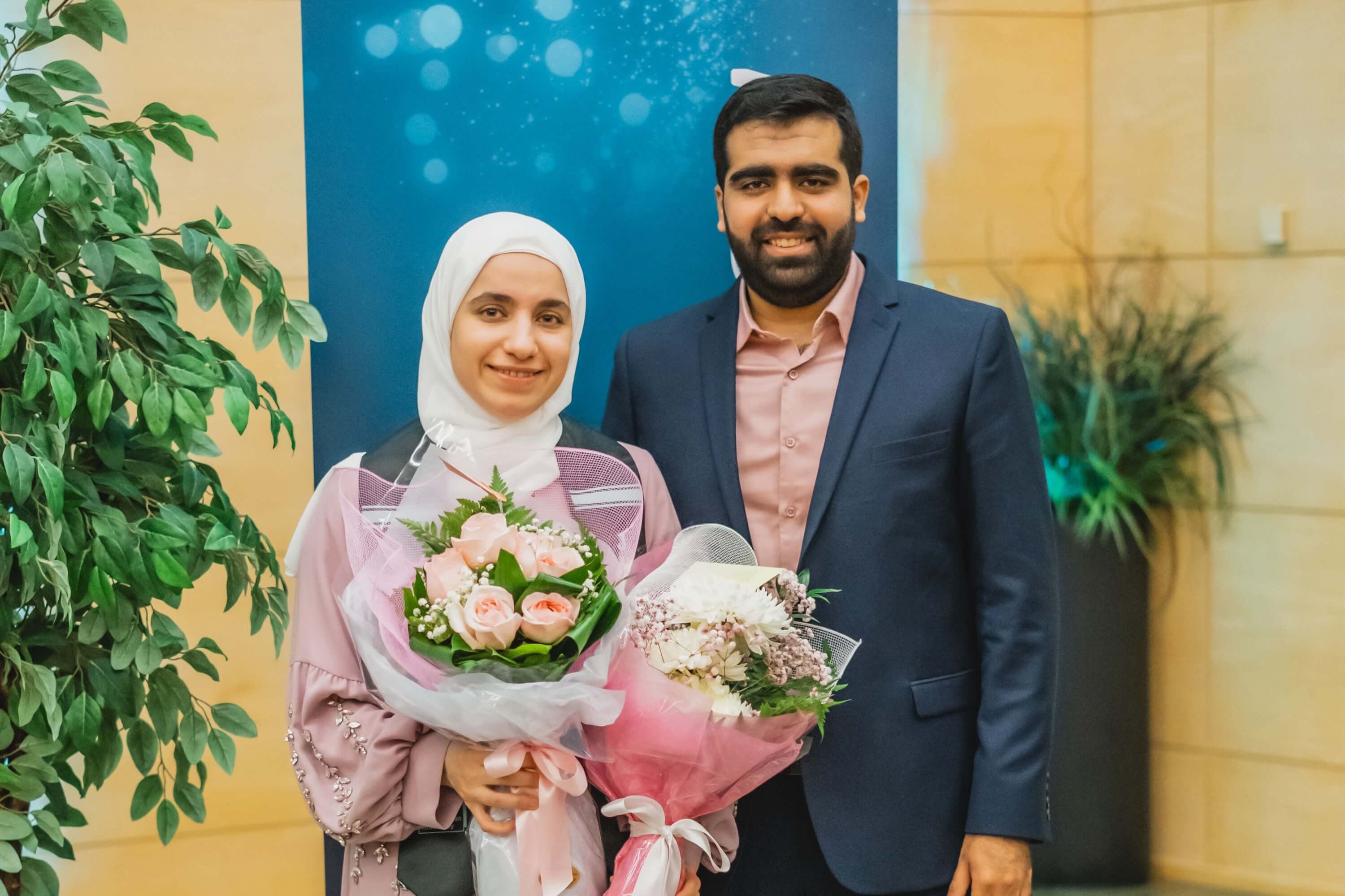 A graduate holding two bouquets of pink flowers standing with a man in a beige shirt and navy blue blazer.