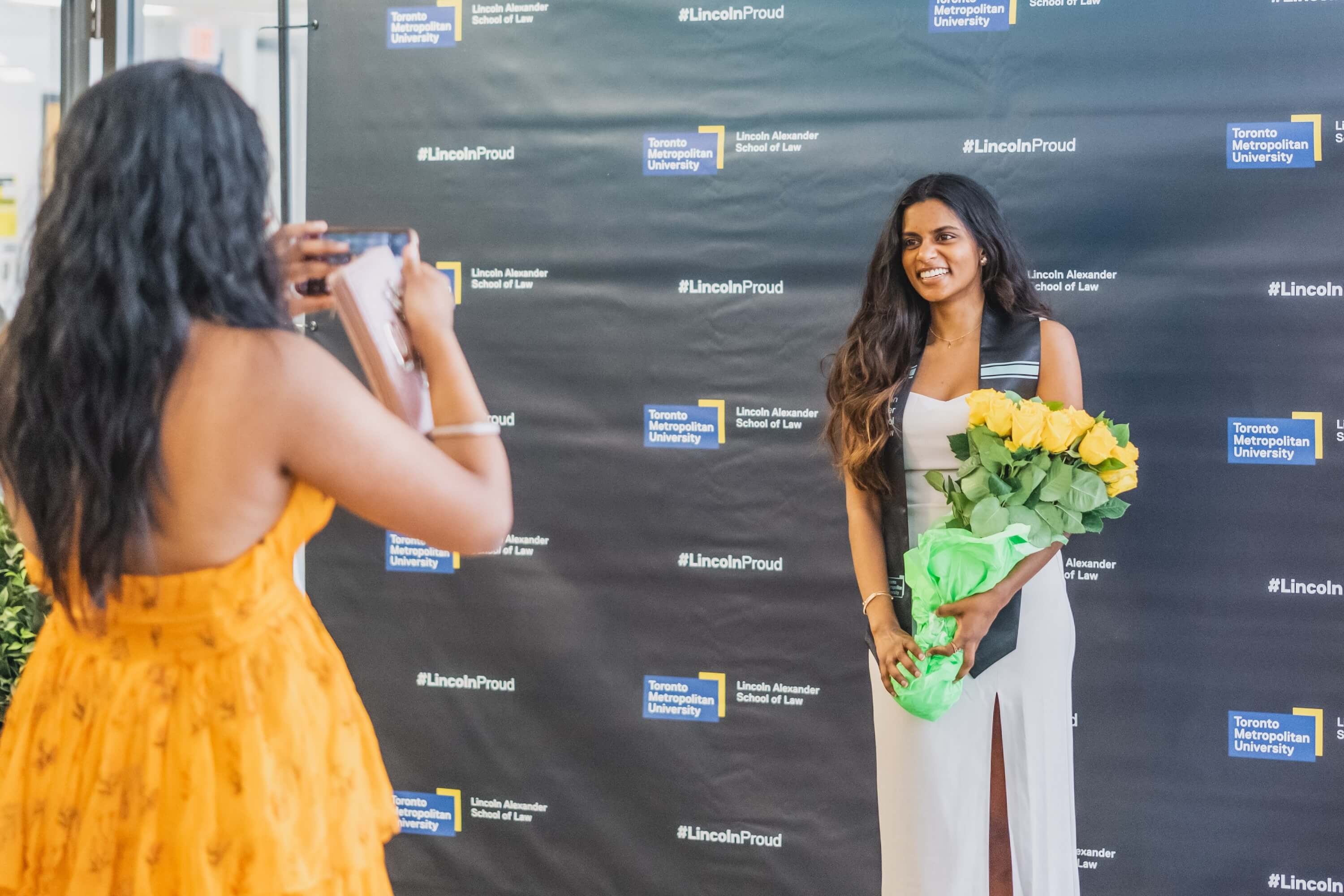 A young woman posing with yellow roses in front of a backdrop while another woman takes her photo.