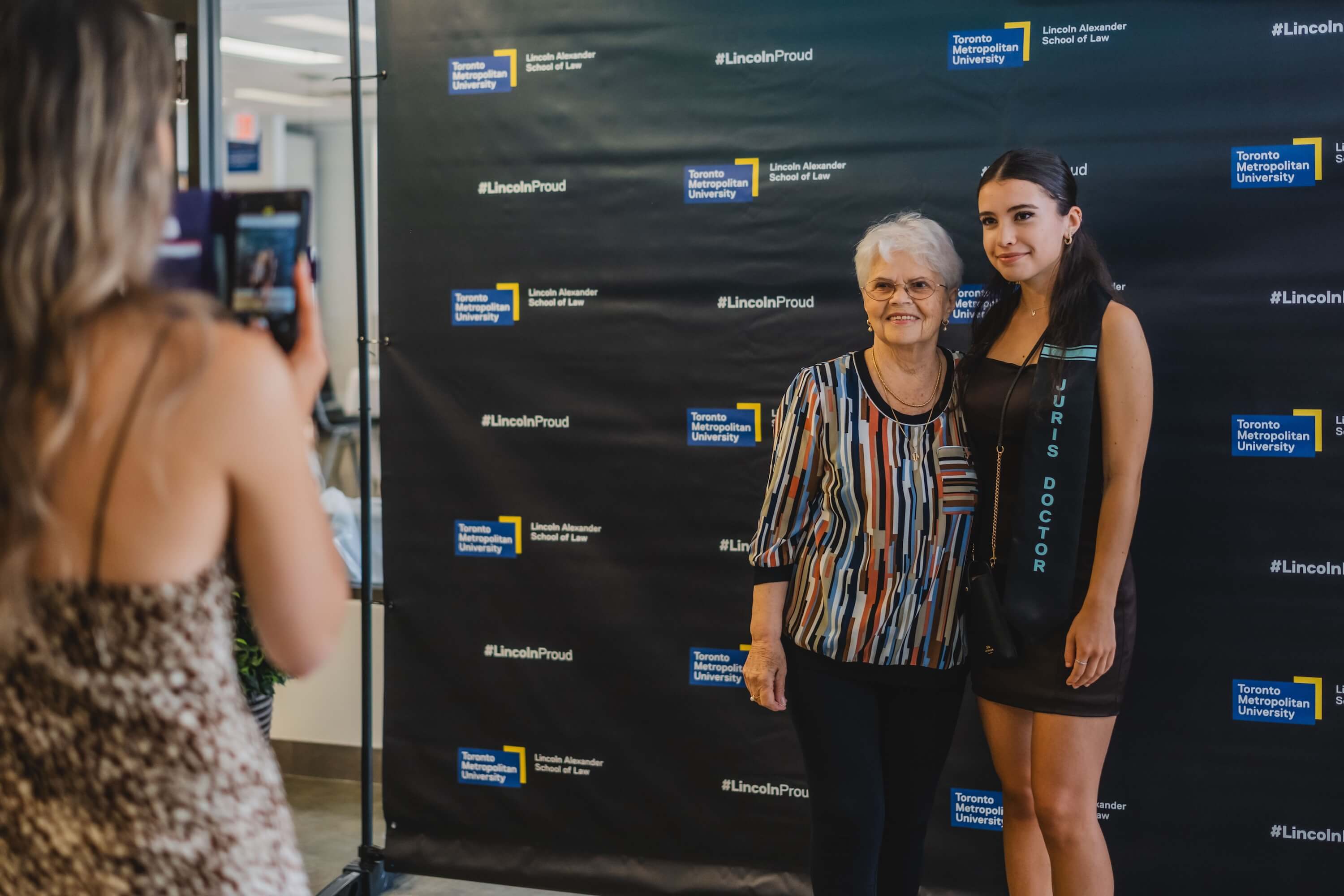 An older woman and a young woman posing in front of a backdrop while a young woman takes their photo.