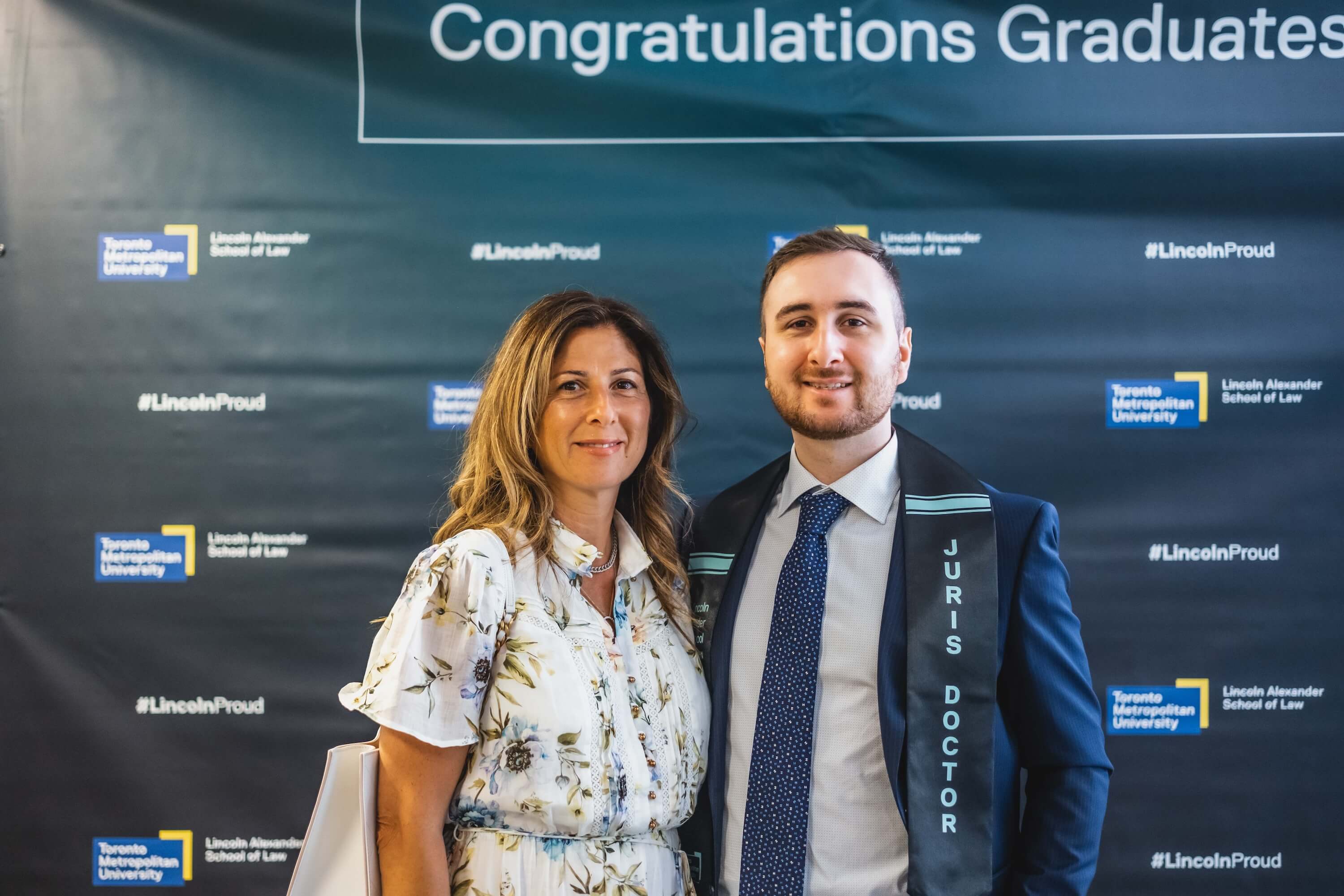 A woman and a young man standing in front of a backdrop that says "Congratulations Graduates!" on it.