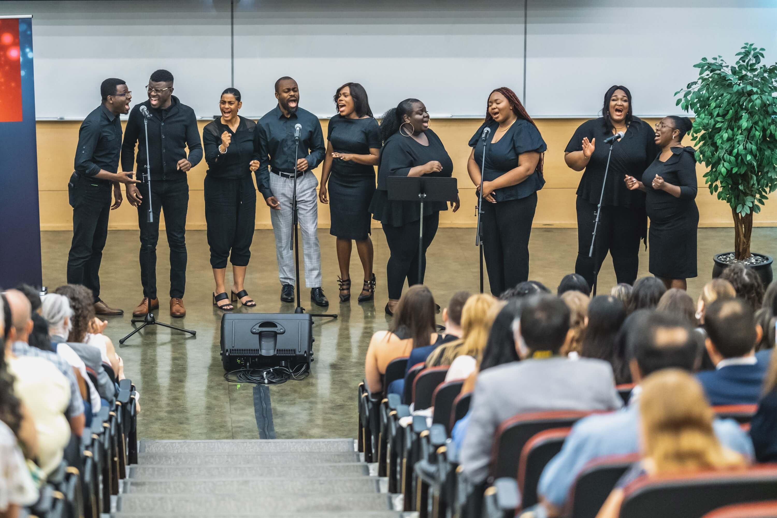 A group of singers dressed in black singing in front of a crowd of people in an auditorium.
