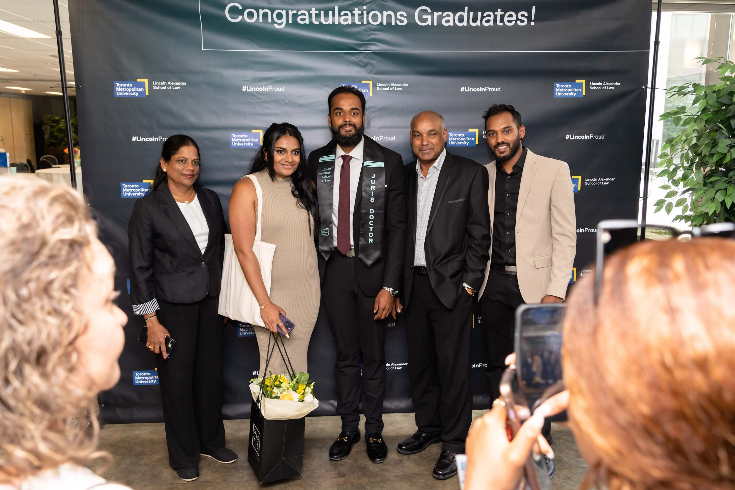 A graduate and four guests pose in front of a backdrop that says "Congratulations Graduates!" while someone takes their photo.