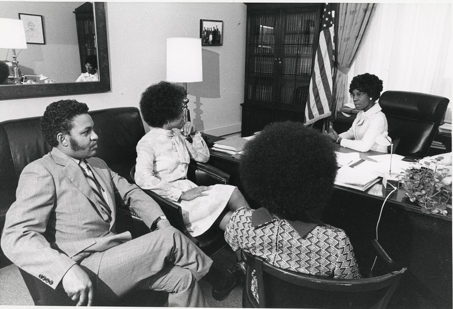 Shirley Chisholm, far right, sitting at desk, talking to three colleagues across from her on the other side of the desk