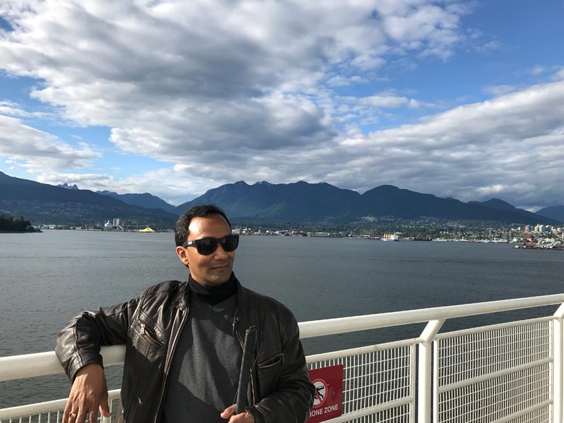 Kevin Shaw, wearing sunglasses, sits outside against a white railing, with a body of water and Vancouver skyline behind him