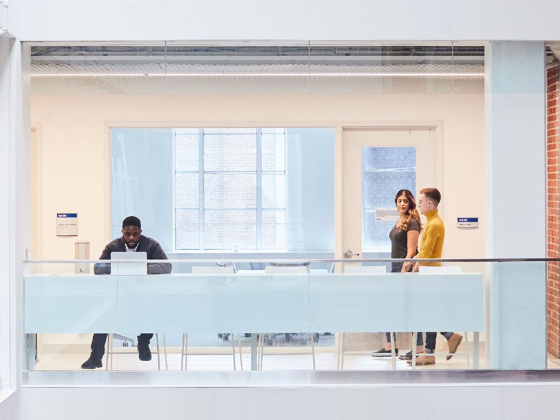 Student on the left sitting at a table working on a laptop while two students on the left walk down the hallway