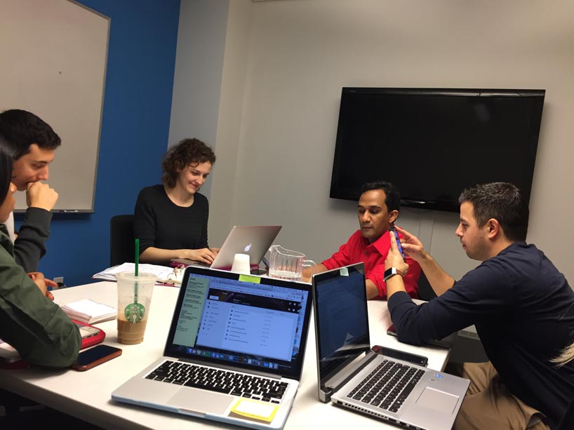 Kevin Shaw, wearing a red shirt, two women and two men wearing dark shirts, sit in a meeting room around a table working on their laptops.]