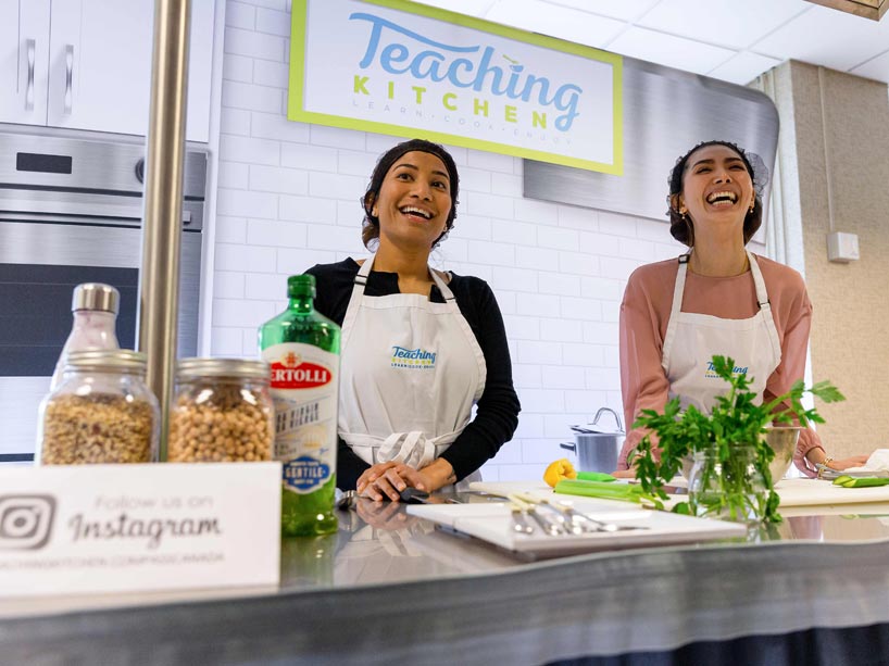 Two women, wearing aprons, laughing behind a kitchen counter