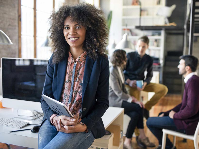 Casually dressed student sits on desk with a group of students collaborating behind her.