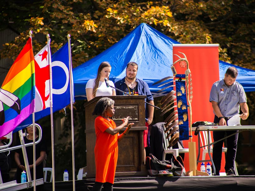 Two students speak at a podium while a man on the right looks down at papers on a table and a woman in front signs ASL