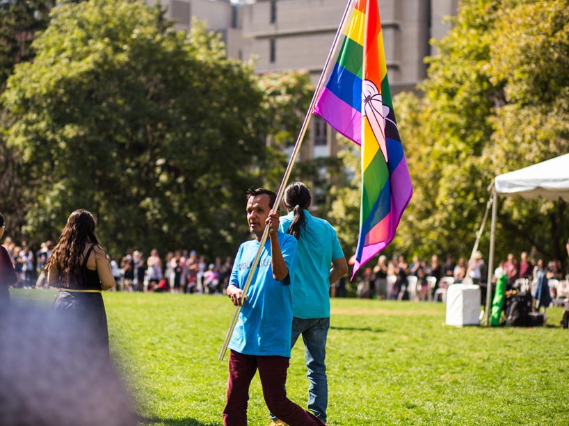 An Indigenous man holds the Two Spirit flag in the quad