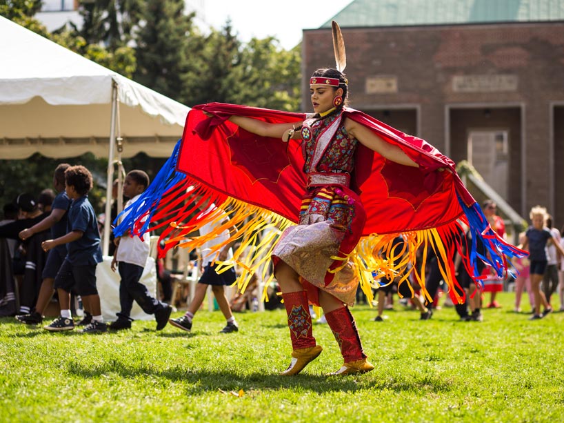 An Indigenous dancer wearing multi-coloured regalia and dancing in the quad