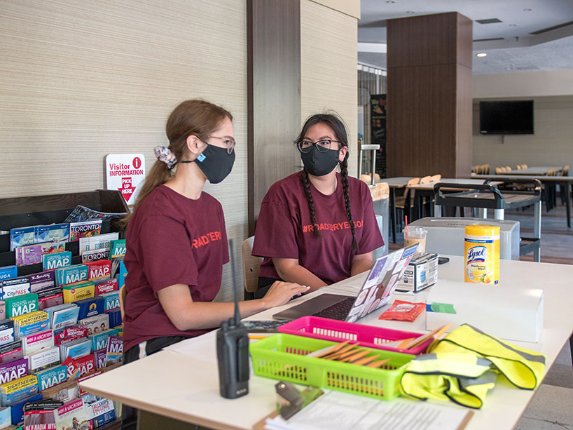 Two young women sit at a desk with face masks on, ready to help students move in at a Ryerson residence