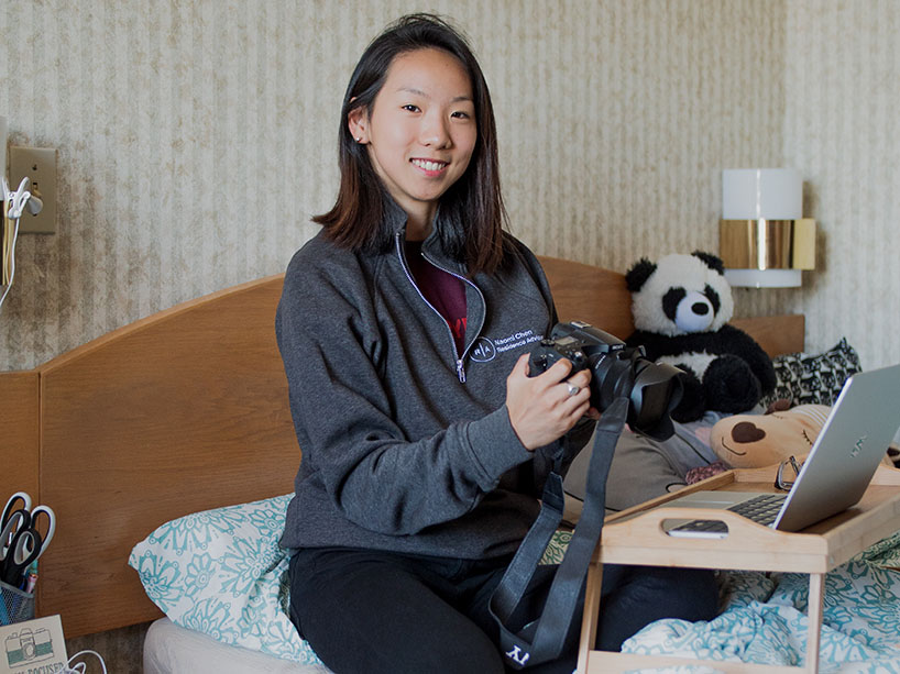 A female student sits on her bed in her room in residence with a camera in hand, smiling