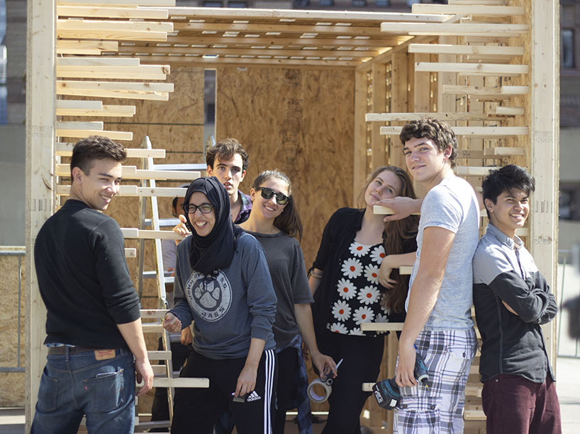 A group of students with building materials pose for a photo in front of a structure they are building