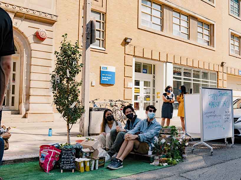 Three people in masks sit on a bench in a mini park