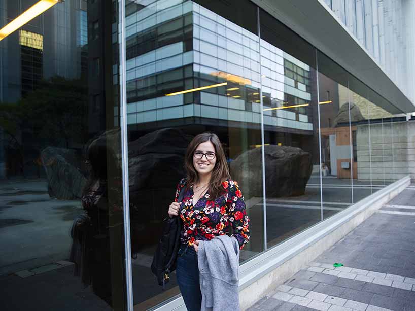 A woman holding a jacket, standing in front of a building in the Ryerson campus with glass windows.