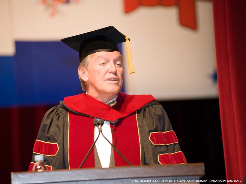 John Honderich in honorary doctorate garb speaks to grads from the podium.