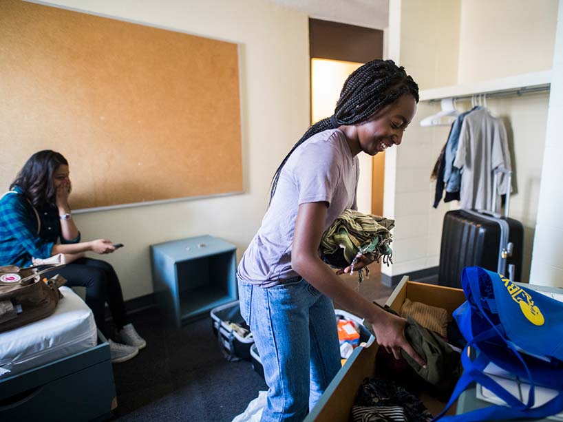 Young woman unpacks clothes into a dresser in her dorm room with a family member behind her.