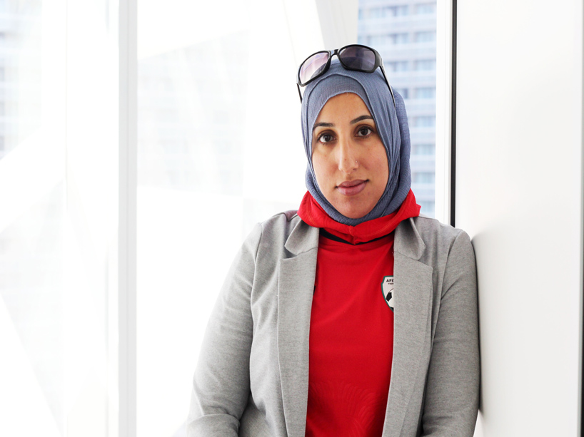 Shireen Ahmed stands against a wall, looking at the camera, with a window behind her. 