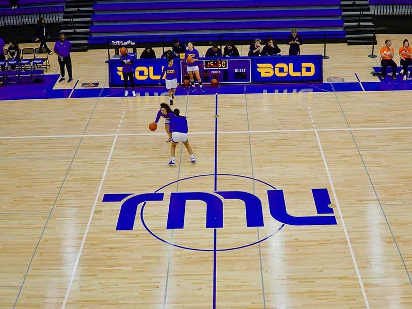 Two people play basketball on the TMU basketball court. A mock-up of the Bold logo is on the sidelines, flanking the time clock.