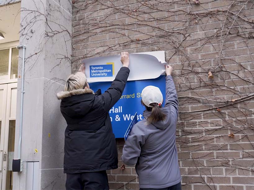 Student affixes a Toronto Metropolitan University branded sign onto the exterior of a building.