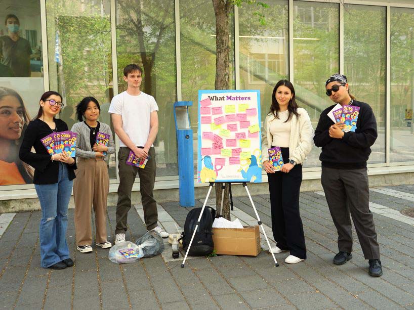 Student volunteers stand with voting booklets and a poster board with sticky notes indicating student priorities.