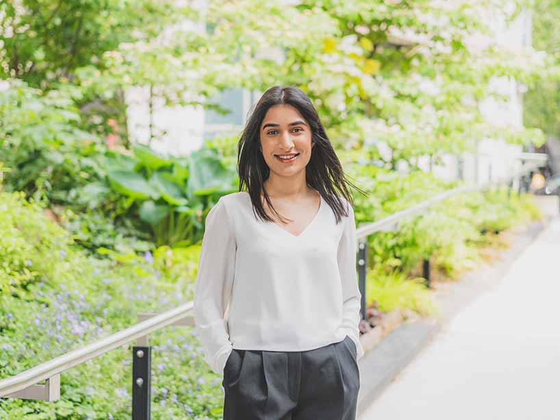 Ridhey Gill in a white blouse and black pants with campus greenery in the background.