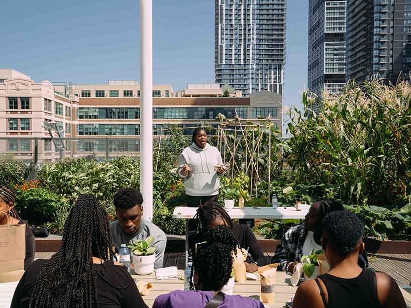 Black woman in grey hoodie speaking to group on Daphne Cockwell Complex rooftop. 