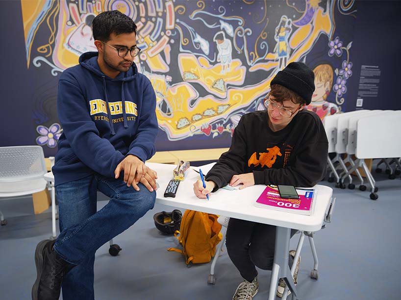 Two students working at a desk in front of the Indigenous wall art.