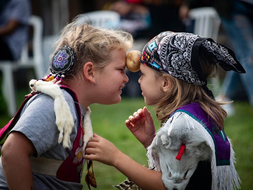 Two youths hold a potato between their foreheads, they are wearing traditional regalia. 