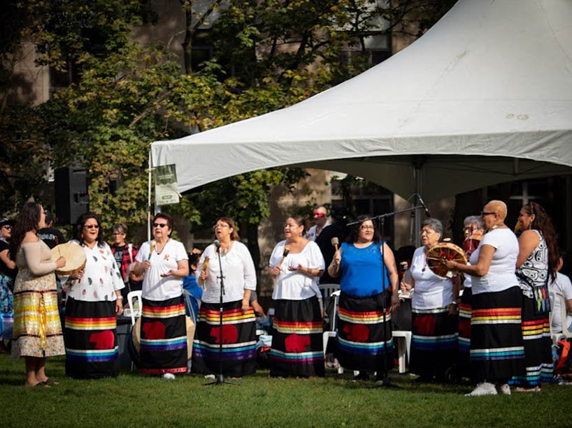 A group of women stand together singing, some play hand drums and others play a shaker. They are dressed in traditional, colourful ribbon skirts. 