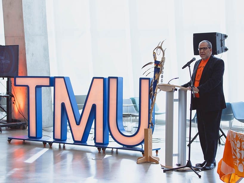 President Lachemi stands at a podium speaking, large TMU letters are beside him.