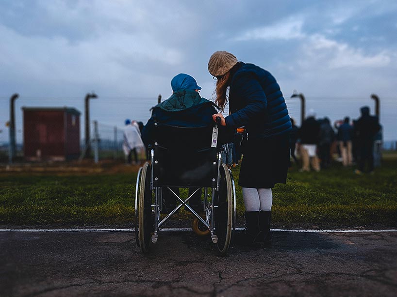 An older man in a wheelchair wearing a cap, with a younger woman standing next to him as both look towards barbed wires at the site of a former concentration camp.