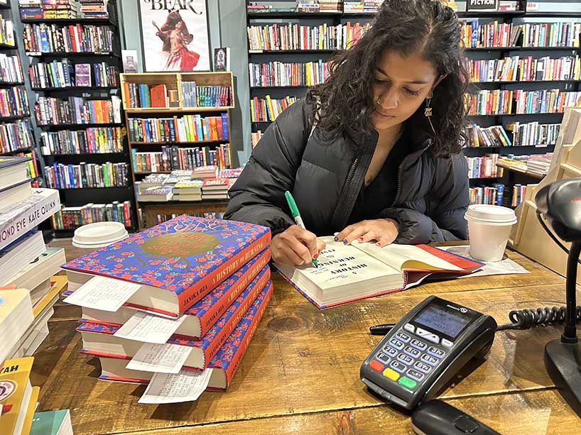 A woman sits at a table signing a stack of books.