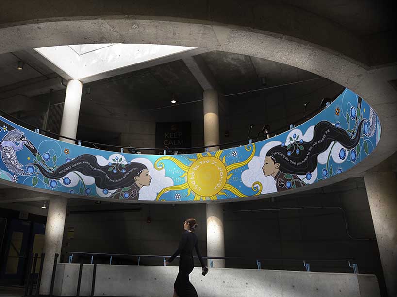 A person walks under the north rotunda of the Recreation and Athletic Centre, above them is an art piece showcasing animals, people and Indigenous symbols
